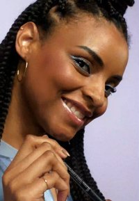Gros plan sur une personne avec des cheveux tressés tenant un crayon eyeliner noir, portant des boucles d'oreilles en or et un haut bleu clair. Fond doux et neutre.