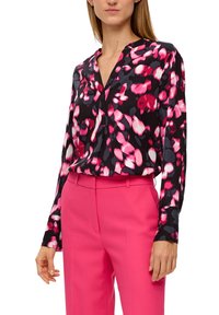 Mujer con blusa de manga larga con un estampado floral abstracto en tonos rosa, blanco y gris, metida dentro de pantalones de talle alto de color rosa brillante.