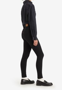 Personne portant un jean noir ajusté avec une couture en forme de cœur sur la poche arrière, une chemise noire boutonnée, des chaussettes blanches et des mocassins noirs.