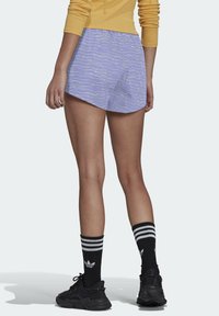 Violet urheilushortsit, joissa on valkoinen aalto-kuvio, kevyestä kankaasta, ja rento istuvuus. Yhdistetty mustiin raidallisiin sukkiin ja tennareihin.