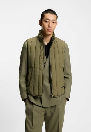 Giovane uomo con capelli corti indossa un gilet imbottito verde oliva con zip sopra un completo coordinato, con le mani in tasca, in piedi davanti a uno sfondo neutro.