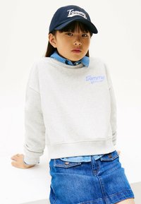 Sudadera gris con el logo "Tommy" en azul claro, llevada sobre una camisa de denim, combinada con una falda de denim. Una gorra navy completa el conjunto.