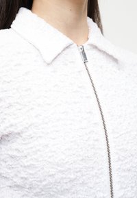 Κοντινό πλάνο ενός ατόμου που φοράει υφασμάτινο λευκό μπουφάν με γιακά και ασημένιο φερμουάρ που τρέχει κάθετα στο εμπρός μέρος.