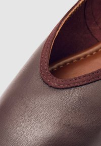 Zapato de cuero marrón con una abertura redonda, que presenta un acabado texturizado y un acento de tela burdeos alrededor del borde de la abertura.