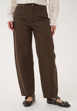 Personne portant un pantalon droit marron, un chemisier en dentelle blanche et des ballerines noires à clous, debout devant un fond clair uni.
