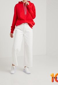 Persoon in een rode hoodie met witte trekkoorden, witte wijde broek en witte sneakers, staande tegen een effen lichte achtergrond.