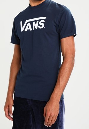 Hombre con una camiseta azul marino de Vans con logo blanco, combinada con pantalones azul oscuro, posando frente a un fondo claro.