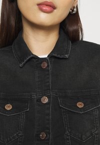 Kvinna som bär en svart denimjacka med metalldekor och stora silverörhängen, som visar hals och nedre ansikte.