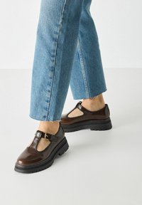 Bruine leren T-band schoenen met een gesp, ronde neus en een gestructureerde zool, gecombineerd met opgerolde blauwe denim jeans.