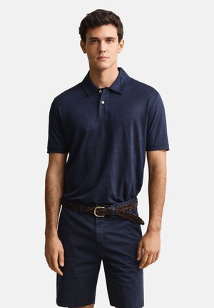Joven con una camiseta polo azul marino, cinturón trenzado marrón y pantalones cortos azul marino, de pie frente a un fondo claro y liso.