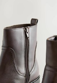 Bottines en cuir marron avec une fermeture éclair sur le côté, présentant une texture lisse, des détails de couture et une petite languette pour un enfilage facile.