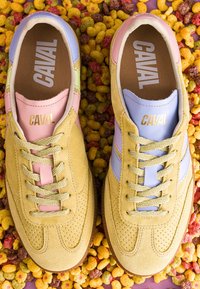 Paire de baskets en daim beige avec des accents pastel rose et bleu et des lacets, étiquetées "CAVAL", posées sur un fond de morceaux de céréales multicolores.
