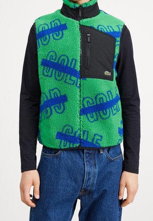Grüne Fleeceweste mit blauen abstrakten Mustern und schwarzer Brusttasche mit Reißverschluss, getragen über einem schwarzen Langarmshirt und blauen Jeans.