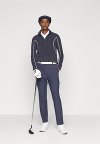 Marineblauer Pullover mit weißen Paspeln, Reißverschlusskragen, kombiniert mit passenden Hosen und weißen Schuhen; hält einen Golfschläger und trägt eine blaue Kappe.