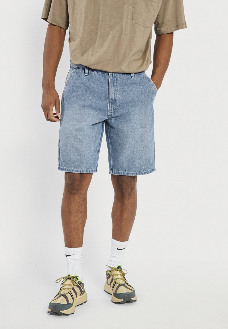 Shorts en denim bleu clair à coupe décontractée, avec passants pour ceinture et une poche unique. Associés à des chaussures de sport de couleur claire et des chaussettes blanches.