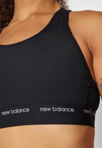 Juodas sportinis liemenukas su besiūliu dizainu, plačiu elastingu diržu su "new balance" logotipu ir prigludusia forma patogumui bei paramai.