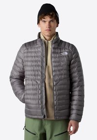 Šedá péřová bunda s horizontálním prošíváním, plný zip, stojáček, nošená přes béžový fleecový top. Černá pletená čepice. Zelené kalhoty.