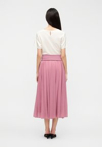 Λευκό τοπ με κοντά μανίκια και άνοιγμα στην πλάτη, σε συνδυασμό με μακριά ροζ πλισέ φούστα, φορεμένα με μαύρα παπούτσια με τακούνι.