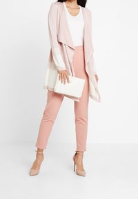 Ljusrosa tygdraperad cardigan med vita randiga detaljer, kombinerad med rosiga byxor och nude-klackar. Håller en vit clutchväska. Mjuk textur.