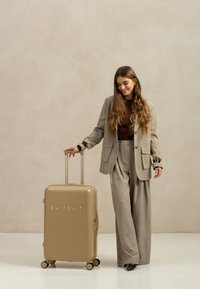 Harde beige koffer met een glanzende afwerking en een uitschuifbare handgreep, uitgerust met vier wielen en het merk "SUITSUIT" in reliëf op de voorkant.