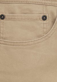 La tela beige presenta un pequeño bolsillo de forma redondeada, con costuras reforzadas y un detalle de botón metálico con un logo grabado.