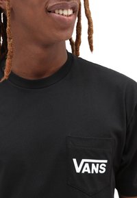 Osoba oblečená v černém tričku Vans s náprsním kapsou, na které je bílé logo Vans, se usmívá a ukazuje část obličeje a vlasů.
