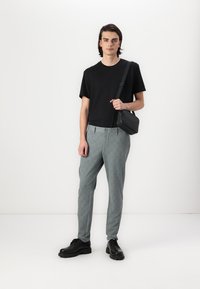 Gray checkered trousers with a flat front, black t-shirt, and black crossbody bag. The model wears black shoes, standing against a white background.