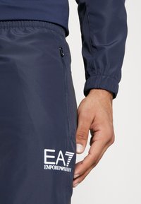 Navy athletic track pants made of smooth fabric, featuring an elastic waistband, zip pocket, and white EA7 branding on the side.