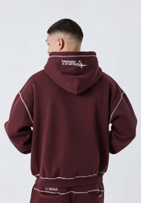 Felpa bordeaux con cappuccio regolabile, caratterizzata da dettagli in cucitura bianca e un logo sul retro. Il tessuto sembra liscio e morbido.
