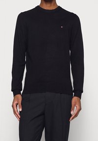 Pull en tricot noir à manches longues, col rond, avec un petit logo sur la poitrine et des poignets et un ourlet côtelés.