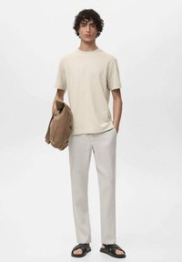 Beige kortærmet t-shirt, lysegrå bukser og sorte sandaler. Modellen holder en tan skuldertaske. Enkel, moderne design med neutrale farver.