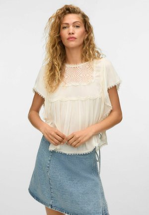 Femme aux cheveux blonds bouclés portant un chemisier en dentelle crème à manches courtes et une jupe en jean bleu clair, debout, mains jointes.