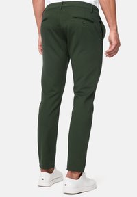 Pantalon vert coupe slim en coton, avec poches arrière à boutonnière et quincaillerie assortie, porté avec des baskets blanches.