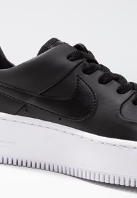 Czarny skórzany sneaker z teksturowaną cholewką, z czarnym logo Swoosh, płaskimi czarnymi sznurowadłami i białą gumową podeszwą z rowkami.