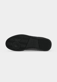 Semelle d'une chaussure noire avec un motif circulaire près du talon, des rainures diagonales au milieu et des crêtes horizontales aux zones des orteils et de la voûte plantaire.