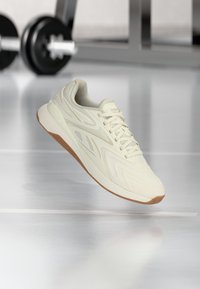 Μπεζ αθλητικό παπούτσι Reebok με καουτσούκ σόλα να αιωρείται πάνω από αντανακλαστικό πάτωμα γυμναστηρίου, με θαμ πή μπάρα άρσης βαρών στο φόντο.