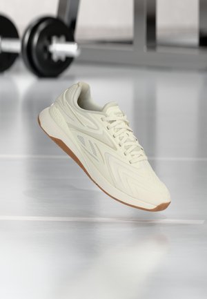 Bež Reebok sportska tenisica s đonom od gume lebdi iznad reflektirajućeg poda teretane, s izmaglicom utega za dizanje u pozadini.