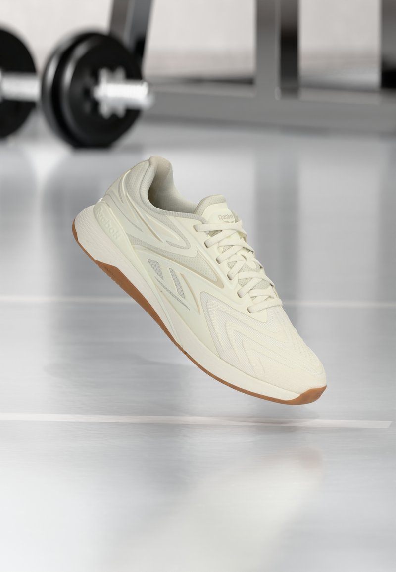 Μπεζ αθλητικό παπούτσι Reebok με καουτσούκ σόλα να αιωρείται πάνω από αντανακλαστικό πάτωμα γυμναστηρίου, με θαμπή μπάρα άρσης βαρών στο φόντο.