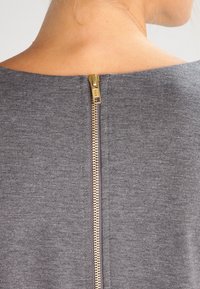 Grå tygplagg med hög halsringning och en gulddragkedja i ryggen, som betonar en mjuk, strukturerad yta.