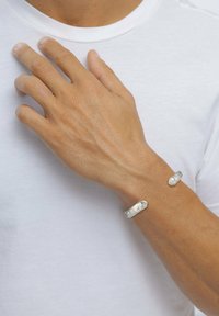 Silverfärgade metallarmband med en texturerad yta, ett tjockare och ett smalare, bärs på en hand mot en enkel vit t-shirt bakgrund.