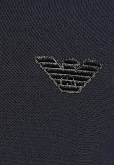 Melna audi ar teksturētu virsmu, kas izceļas ar izšūtu logotipu, kas veidots spārnu formā, virs burtiem "EA" ir horizontālas svītras.