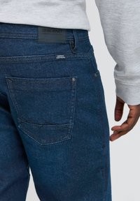 Jeans de mezclilla azul oscuro con un acabado texturizado, que presentan un parche de logo negro en la cintura y costuras en contraste en el bolsillo trasero.