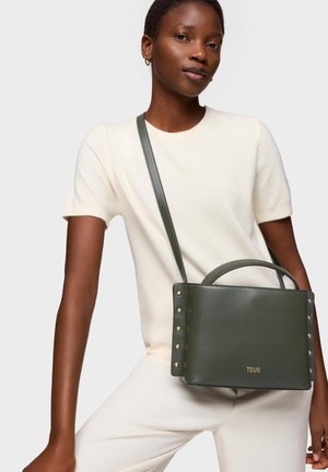 Mujer con top de manga corta color crema y pantalones, llevando un bolso cruzado de cuero verde oscuro con tachuelas doradas y logo.