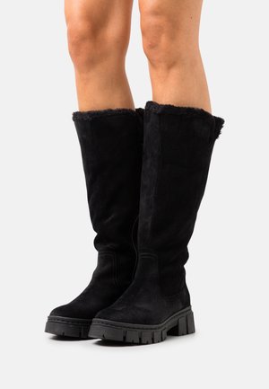 Bottes noires montantes jusqu'aux genoux avec semelles en caoutchouc épaisses et doublure en fourrure portées sur des jambes nues sur un fond clair uni.