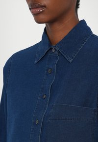 Gros plan d'une personne portant une chemise en denim bleu foncé à boutons, avec un col et une poche poitrine, montrant son cou et le bas de son visage.