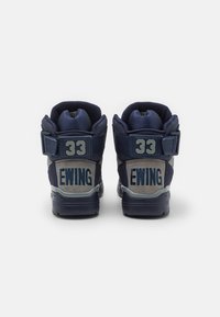 Paire de baskets montantes bleu marine et gris avec "EWING" et le numéro "33" sur les lanières arrière, présentées de dos sur un fond blanc.