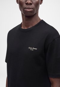 Gros plan d'une personne portant un T-shirt noir avec le texte blanc "Samsøe Samsøe Since 1993" sur le côté gauche de la poitrine.