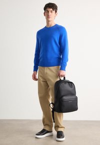 Maglione blu a maglia, pantaloni beige, zaino in pelle nera con tasca frontale, e sneakers nere con suola bianca, su uno sfondo neutro.
