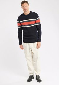 Homme portant un pull marine avec des rayures rouges et blanches horizontales, un pantalon couleur crème et des bottes noires, se tenant devant un fond blanc uni.