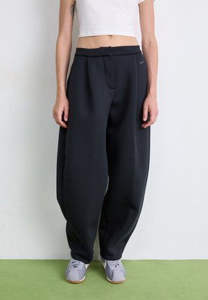 Personne portant un pantalon noir ample, un crop top blanc et des baskets grises, debout contre un mur blanc sur un sol vert.